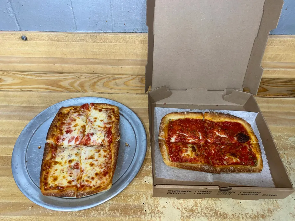 Two slices of cheese pizza on a metal tray, and another two slices of cheese pizza in a cardboard pizza box on a wooden table.