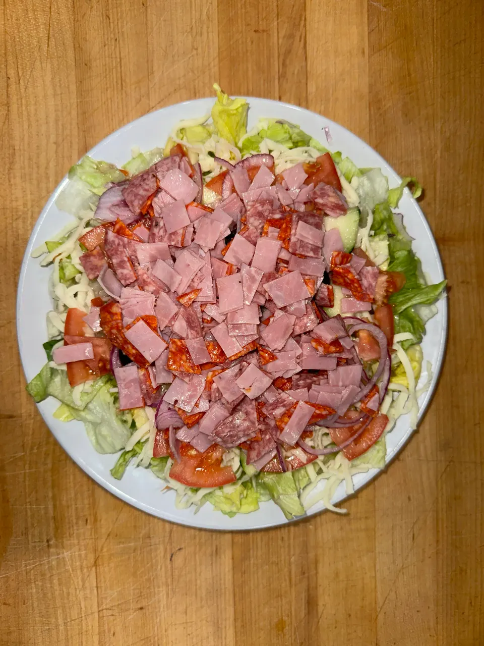 Plate of salad with lettuce, tomatoes, onions, shredded cheese, and chopped ham on top, placed on a wooden surface.